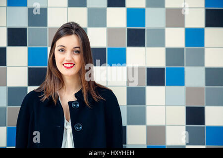 Attraktive junge Geschäftsfrau oder Student mit verschränkten Armen gegen eine blaue Mosaikwand lächelnd Stockfoto
