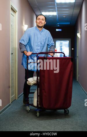 Junger Mann schob einen Haushalt Wagen beladen mit saubere Handtücher, Wäsche und Reinigungsgeräte in einem Hotel, als er das Zimmer services Stockfoto