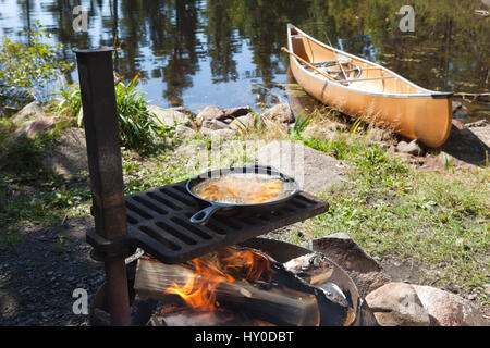 Fisch kochen in einer Pfanne über dem offenen Feuer mit einem Kanu und nördlichen Minnesota See im Hintergrund Stockfoto