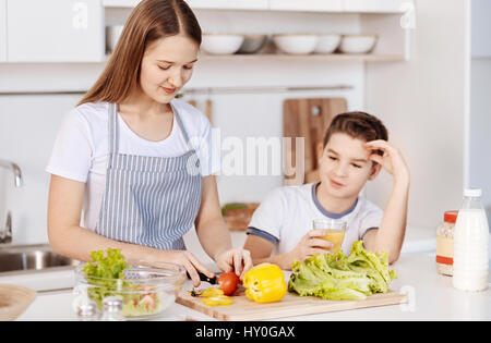 Gesunde Ernährung. Positiv erfreut Teanage Mädchen Schneiden von Gemüse und Salat stehen in der Küche mit ihrem jüngeren Bruder vorbereiten Stockfoto