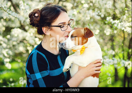 Junge schöne Frau küsst ihren geliebten Hund Jack Russell Terrier auf einem Hintergrund von Frühjahr blühende Bäume Stockfoto