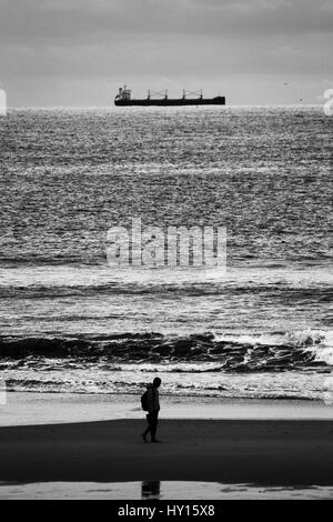 Ein einsamer Mann zu Fuß auf dem Sand des Strandes von Matosinhos, während ein Schiff entlang der Küste navigiert. Stockfoto