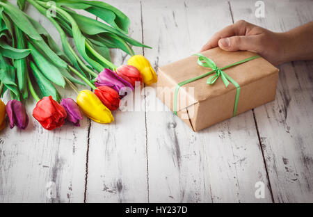Blumenstrauß lila, rote und gelbe Tulpe und Mannes Hand hält eine Geschenkbox auf hölzernen Hintergrund. Geburtstag, Muttertag oder Frau Grüße Stockfoto