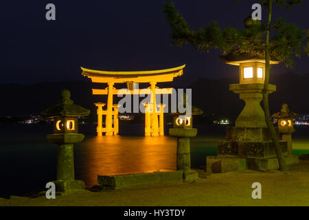 Beleuchtete schwimmende Torii-Tor im Hintergrund in der Nacht mit beleuchteten Steinlaternen im Vordergrund in Miyajima, Japan Stockfoto