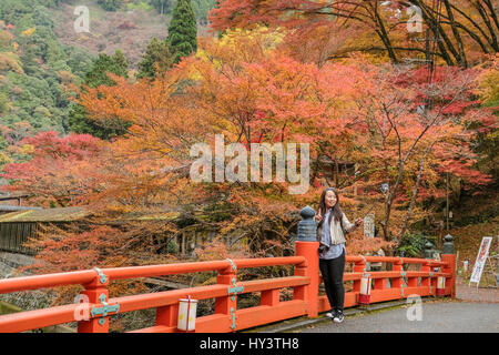 Frau steht auf rote Brücke Herbst Farbe Bäumen umgeben und nimmt Selfie Foto mit Handy im Jingoji-Tempel in Kyoto, Japan Stockfoto