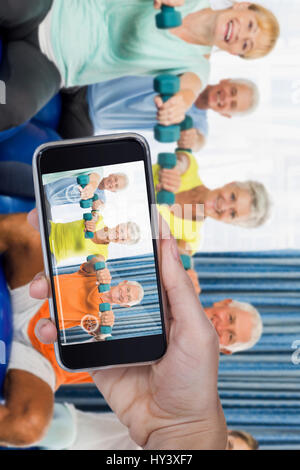 Hand halten Handys vor weißem Hintergrund gegen Porträt von Senioren mit Gymnastikball und Gewichte Stockfoto