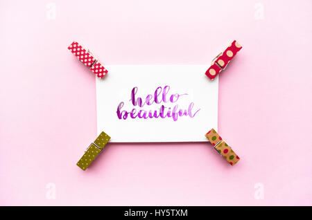 Inspirierende Zitat Hallo schön handschriftlich mit Aquarell in Kalligraphie-Stil, Mini Wäscheklammern auf einem rosa Hintergrund. Flach zu legen. Stockfoto