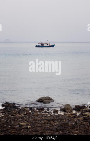 Ein Taucher Transport Boot vor der Küste an einem ruhigen langweiligen grauen Tag mit Felsstrand Vordergrund verankert. Stockfoto
