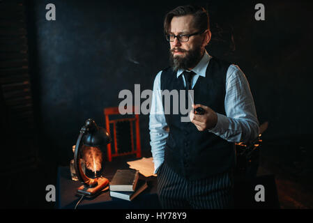 Ernsthafte bärtigen Schriftsteller in Gläsern raucht eine Pfeife. Feder, Kristallkaraffe, Bücher und Vintage Lampe auf dem Schreibtisch Stockfoto