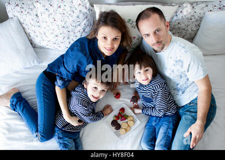 Schöne Familie von vier, liegend auf dem Bett, Essen Erdbeeren und Cookies, Blick in die Kamera, geschossen von oben, Mutter, schwanger zu sein Stockfoto