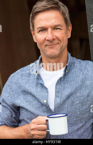 Porträt gedreht von einem attraktiven, erfolgreichen und glücklichen Mann mittleren Alters männlichen trägt ein blaues Hemd trinken Tee oder Kaffee aus einer Blechtasse Stockfoto
