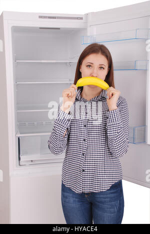 Ein junges Mädchen hält eine Banane in den Mund, ein Lächeln zu imitieren. Schöne junge Mädchen in der Nähe der Kühlschrank. Stockfoto