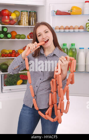 Junges Mädchen hält Würstchen auf dem Kühlschrank Hintergrund. Schöne junge Mädchen in der Nähe der Kühlschrank. Stockfoto