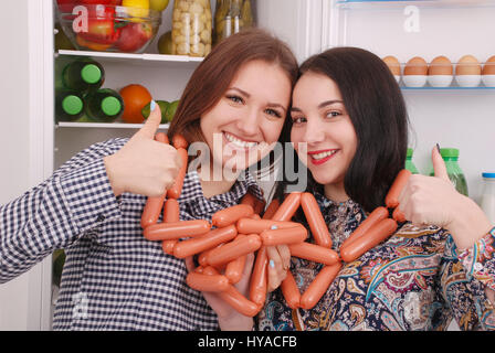 Zwei junge Mädchen hält Würstchen auf dem Kühlschrank Hintergrund. Zwei schöne junge Mädchen in der Nähe der Kühlschrank. Stockfoto