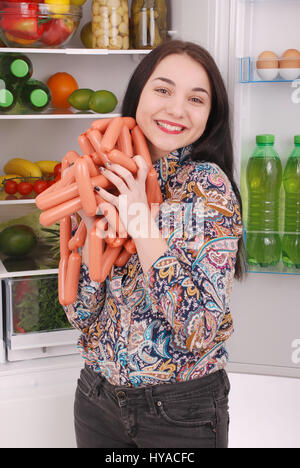 Junges Mädchen hält Würstchen auf dem Kühlschrank Hintergrund. Schöne junge Mädchen in der Nähe der Kühlschrank. Stockfoto