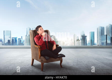 Durchdachte asiatische Geschäftsmann sitzt auf dem Sofa denken über etwas mit Wolkenkratzer Hintergrund Stockfoto