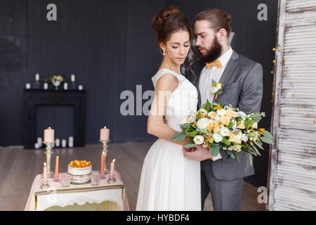 Die Braut mit einem Blumenstrauß in der Hand und der Bräutigam stehen nebeneinander in einem schönen Interieur. Die Braut im Mittelpunkt. Stockfoto