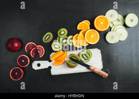 Frische Orangen, Kiwi und Äpfel werden in Scheiben geschnitten und mit Messer auf dunklen grauen Oberfläche, Top Aussicht mit weißen Holzbrett gelegt Stockfoto
