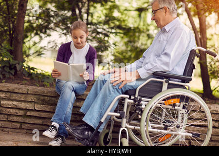 Lesen Buch im freien fröhlich Enkelin Zeit miteinander zu verbringen und deaktiviert Großvater im Rollstuhl Stockfoto