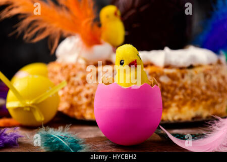 Nahaufnahme von einem Spielzeug-Küken aus einem rosa Ei, einige Ostereier und Federn in verschiedenen Farben auf einer Holzfläche und ein Mona de Pascua, ein Kuchen-ea Stockfoto
