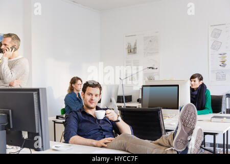 Mitte erwachsener Mann im Büro, Füße auf Schreibtisch Teetasse Blick in die Kamera halten Stockfoto