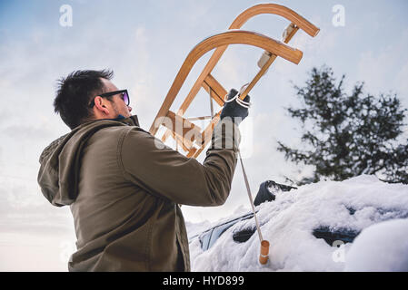 Mann trägt graue Winterjacke, Schlitten auf Autodach Stockfoto