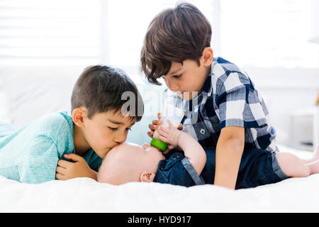 Ältere Brüder (6-7, 8-9) füttern kleinen Bruder (12-17 Monate) liegen auf dem Bett Stockfoto