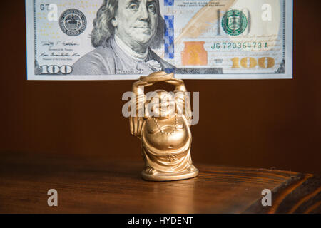 eine kleine goldene Buddha Figur stehend auf einem Holztisch hält einen hundert-Dollar-Schein in Vereinigte Staaten Währung über seinem Kopf Stockfoto