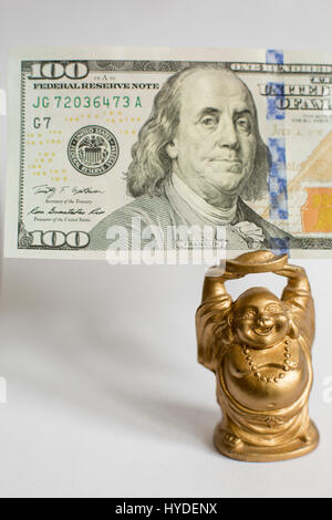 eine kleine goldene Buddha Figur stehend auf einem weißen Hintergrund hält einen hundert-Dollar-Schein in Vereinigte Staaten Währung über seinem Kopf Stockfoto