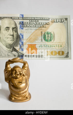 eine kleine goldene Buddha Figur stehend auf einem weißen Hintergrund hält einen hundert-Dollar-Schein in Vereinigte Staaten Währung über seinem Kopf Stockfoto