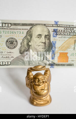 eine kleine goldene Buddha Figur stehend auf einem weißen Hintergrund hält einen hundert-Dollar-Schein in Vereinigte Staaten Währung über seinem Kopf Stockfoto