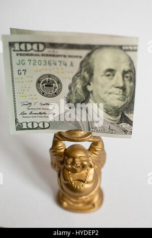 eine kleine goldene Buddha Figur stehend auf einem weißen Hintergrund hält einen hundert-Dollar-Schein in Vereinigte Staaten Währung über seinem Kopf Stockfoto