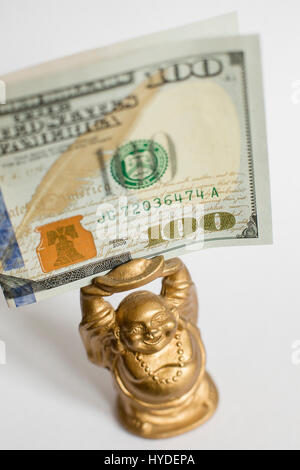 eine kleine goldene Buddha Figur stehend auf einem weißen Hintergrund hält einen hundert-Dollar-Schein in Vereinigte Staaten Währung über seinem Kopf Stockfoto