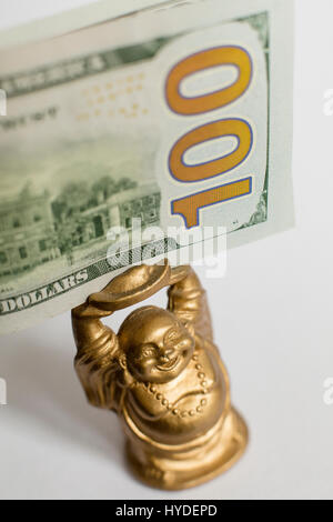 eine kleine goldene Buddha Figur stehend auf einem weißen Hintergrund hält einen hundert-Dollar-Schein in Vereinigte Staaten Währung über seinem Kopf Stockfoto