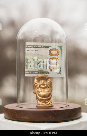 eine kleine goldene Buddha Figur stehend in einem Glas Kuppel hält ein gefalteten hundert Dollar bill über seinem Kopf angezeigt Stockfoto