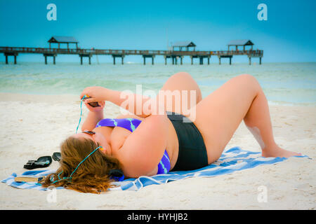 Übergrößen Frau in Zweiteiliger Badeanzug am Strand von Clearwater, FL holding Smart Phone mit Ohrhörer und Pier 60 im Golf von Mexiko liegen Stockfoto