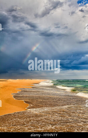 Regenbogen im hellen Strahlen des Lichts bei stürmischen Regenwetter über dem Sandstrand und pazifischen Ozeanwellen Surfen in Stockton Beach, New South Wales, Australien. Stockfoto