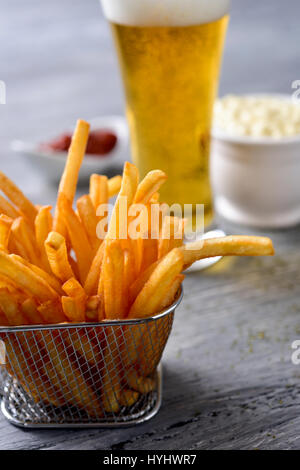 Nahaufnahme von einigen leckeren Pommes frites in einen Metallkorb auf einem grauen rustikalen Holztisch und ein Glas mit Bier und einige Schüsseln mit Mayonnaise serviert ein Stockfoto