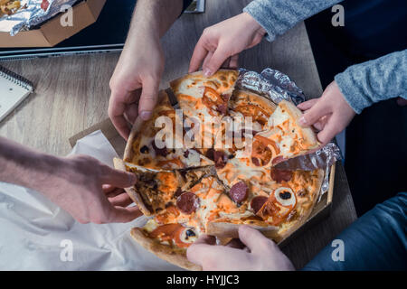 Pizza essen gemeinsam im Büro. Draufsicht der Hände unter Pizza. Begriff der Freundschaft am Arbeitsplatz, Einheit, Teamarbeit, Partnerschaft. Stockfoto