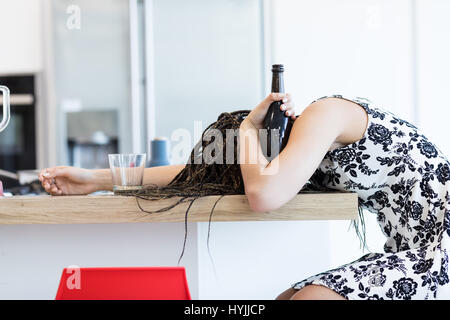 Seitenansicht der betrunkene junge Frau mit Zöpfen am Tisch verdeckt liegen und eine Flasche Weine in der Hand hält, über weiße Küche Hintergrund Stockfoto