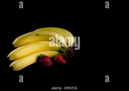 Vier frische goldene Bananen mit zwei Erdbeeren in dunklem Hintergrund isoliert Stockfoto