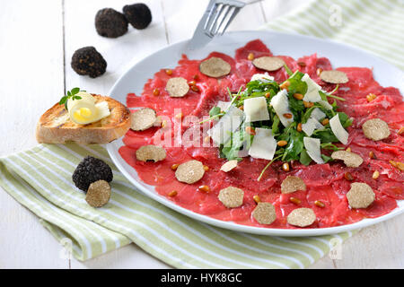 Köstliches Rindfleisch-Carpaccio mit in Scheiben geschnittenen schwarzen Trüffeln, Rucola-Salat mit Parmesan und gerösteten Pinienkernen, serviert mit einer Scheibe geröstetes baguette Stockfoto