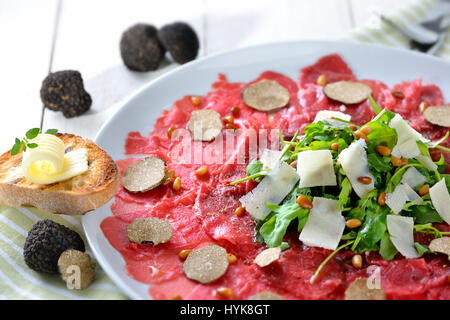 Köstliches Rindfleisch-Carpaccio mit in Scheiben geschnittenen schwarzen Trüffeln, Rucola-Salat mit Parmesan und gerösteten Pinienkernen, serviert mit einer Scheibe geröstetes baguette Stockfoto