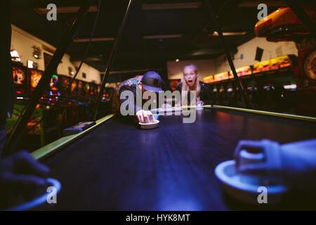 Glückliche junge Freunde spielen Luft-Hockey-Spiel im Freizeitpark. Mann trifft Puck mit Stürmer. Stockfoto