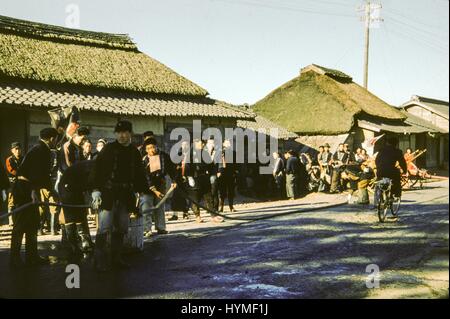 Eine große Gruppe von japanischen Männer in uniform, mögliche Feuerwehrleute halten einen Schlauch, der auf der Straße eines traditionellen Dorfes erstreckt sich wie ein Mann vorbei fährt mit dem Fahrrad, Japan, 1952. Stockfoto