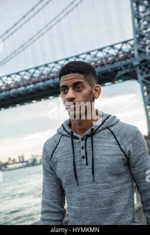 Ein Porträt von einem jungen, schwarzen Mann, sinnend stehen entlang der New Yorker East River und Williamsburg Bridge. Stockfoto
