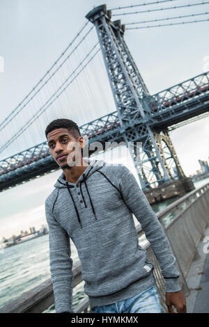 Ein junger Mann wartet entlang der East River, darunter die Williamsburg Bridge. Während des Sonnenuntergangs ein Herbst in New York City gedreht. Stockfoto