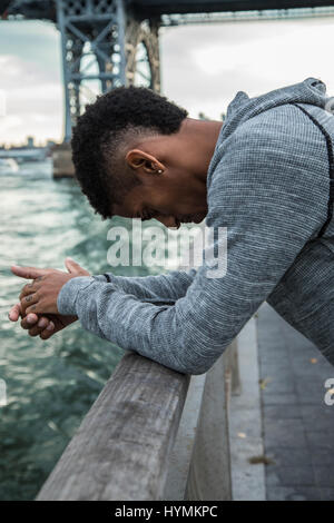 Ein Porträt von einem jungen, schwarzen Mann, sinnend stehen entlang der New Yorker East River und Williamsburg Bridge. Stockfoto