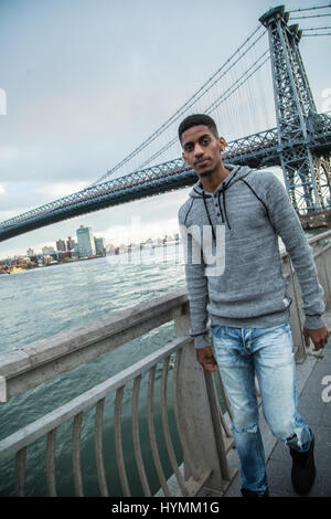 Ein junger Mann wartet entlang der East River, darunter die Williamsburg Bridge. Während des Sonnenuntergangs ein Herbst in New York City gedreht. Stockfoto