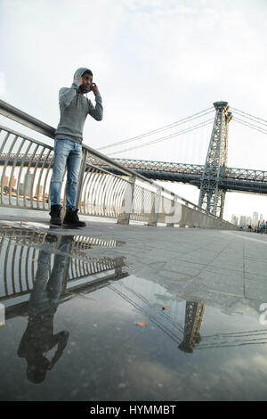 Ein junger Mann wartet entlang der East River, darunter die Williamsburg Bridge. Während des Sonnenuntergangs ein Herbst in New York City gedreht. Stockfoto